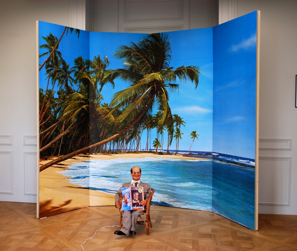 Monsieur Teste (1975), mannequin automate assis sur une chaise en osier, journal,  photo de plage et palmiers, Estate Marcel Broodthaers. Salle 9.